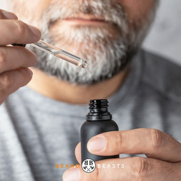 A close-up photo of a man with a salt-and-pepper beard, holding a dropper filled with clear liquid above an open matte black bottle labeled with the Beard Beasts logo.