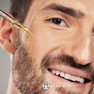 Close-up of a smiling man applying beard oil with a dropper, highlighting the benefits of using the best oils for beard growth, for Beard & Beasts article.