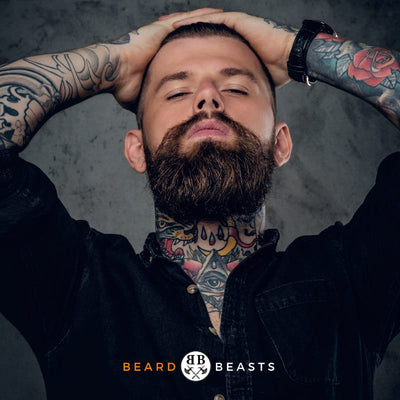 Portrait of a tattooed man with hands behind his head, showcasing a thick, tamed beard, embodying the style promoted by 'Beard Beasts' brand. The image exemplifies the polished look achievable with tips on 'how to tame your beard'.
