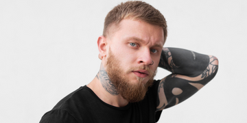 Stylish man with a short fade haircut, beard, and tattoos posing with hand on head. Showcasing a trendy and clean-cut look, ideal for 2024 hairstyles
