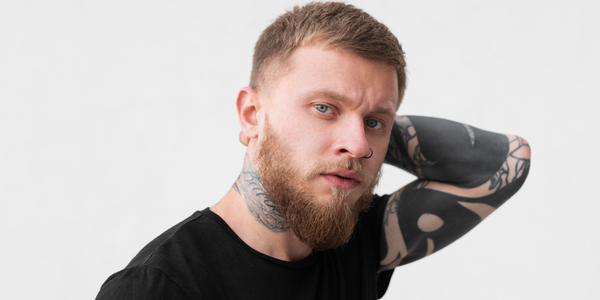 Stylish man with a short fade haircut, beard, and tattoos posing with hand on head. Showcasing a trendy and clean-cut look, ideal for 2025 hairstyles