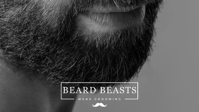 A close-up black and white image focusing on a thick, textured beard on a man's face, with a partial view of his smiling mouth, accompanied by the 'BEARD BEASTS - MEN'S GROOMING' logo, possibly for an article discussing 