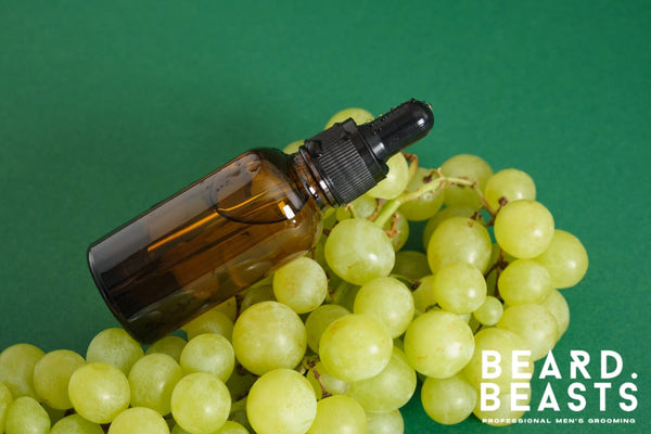 Amber dropper bottle of grapeseed oil for beard care resting on a cluster of fresh green grapes, set against a vibrant green background.