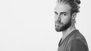 Man with a 2 month beard and man bun hairstyle looking to the side against a white background.