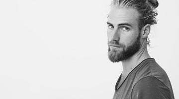 Man with a 2 month beard and man bun hairstyle looking to the side against a white background.