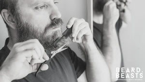 Man trimming his beard with scissors and comb in front of a mirror, demonstrating beard trimming tips for a sharp look