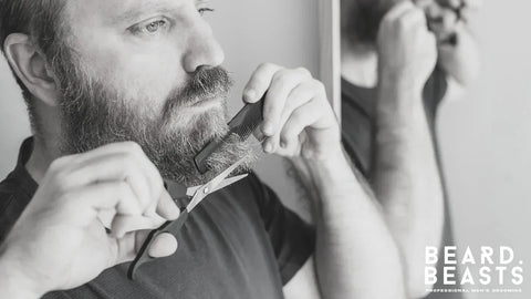 Man trimming his beard with scissors and comb in front of a mirror, demonstrating beard trimming tips for a sharp look