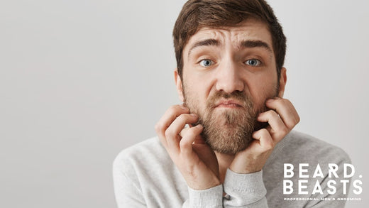 Man with a full beard wearing a light gray shirt, holding his beard with both hands and showing discomfort, likely due to beard rash.