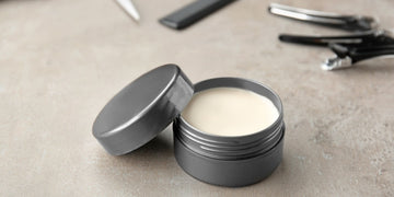 Open container of light-colored pomade with its gray lid off, set on a textured gray surface alongside hair styling tools, illustrating how to use pomade on short hair