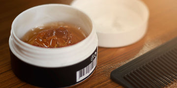 Open container of amber-colored hair pomade next to a white container and black comb on a wooden surface, illustrating essential styling products for modern pomade hairstyles.