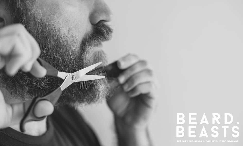 Close-up of a man trimming his beard with scissors—featured image for how to trim a beard while growing it out.