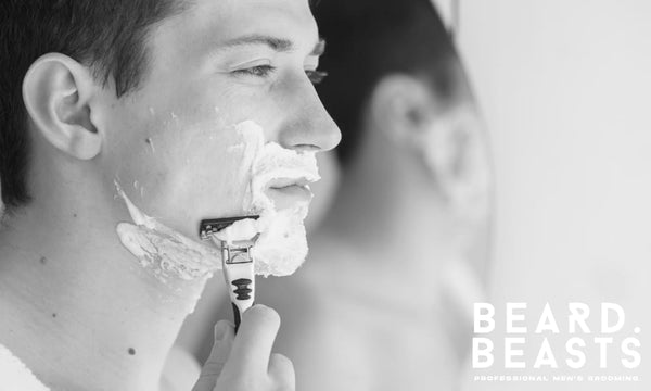 Man demonstrating what is wet shaving with razor and lather on face, seen in mirror reflection during grooming routine.