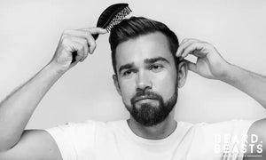 Bearded man brushing his styled hair after applying matte clay, wearing a white t-shirt against a plain background.