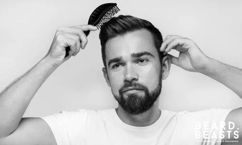 Bearded man brushing his styled hair after applying matte clay, wearing a white t-shirt against a plain background.