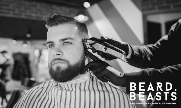A black-and-white photograph of a bearded man sitting in a barbershop chair while a barber uses clippers to shape his low taper fade haircut. 