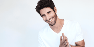 A cheerful man with a 5mm beard, styled and well-groomed. He has curly dark hair, a bright smile, and is wearing a plain white V-neck t-shirt. The background is light and minimal, focusing the attention on his friendly expression.