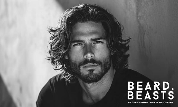 Man with thick, shoulder-length wavy hair and subtle layers, sitting against a sunlit wall—showcasing a natural and classic option for men’s long haircuts for thick hair.