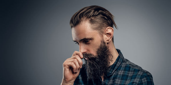 Man with a stylish slicked back haircut and a full beard, wearing a plaid shirt, looking thoughtful. Ideal example of a modern, sophisticated slicked back hairstyle.