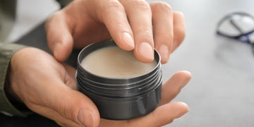 Close-up of a man's fingers scooping pomade from a black tin, demonstrating how to use pomade on curly hair with a focus on the product's creamy texture.