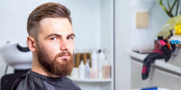Man with an undercut fade hairstyle and full beard in a barber shop, showcasing the undercut vs taper contrast with long, slicked-back top and short, faded sides.